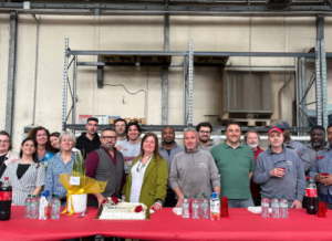 Group photo of about 18 smiling colleagues posing in a warehouse behind a red-covered table with drinks, a decorated cake reading “MLT Cuccolini”. Company celebration scene.