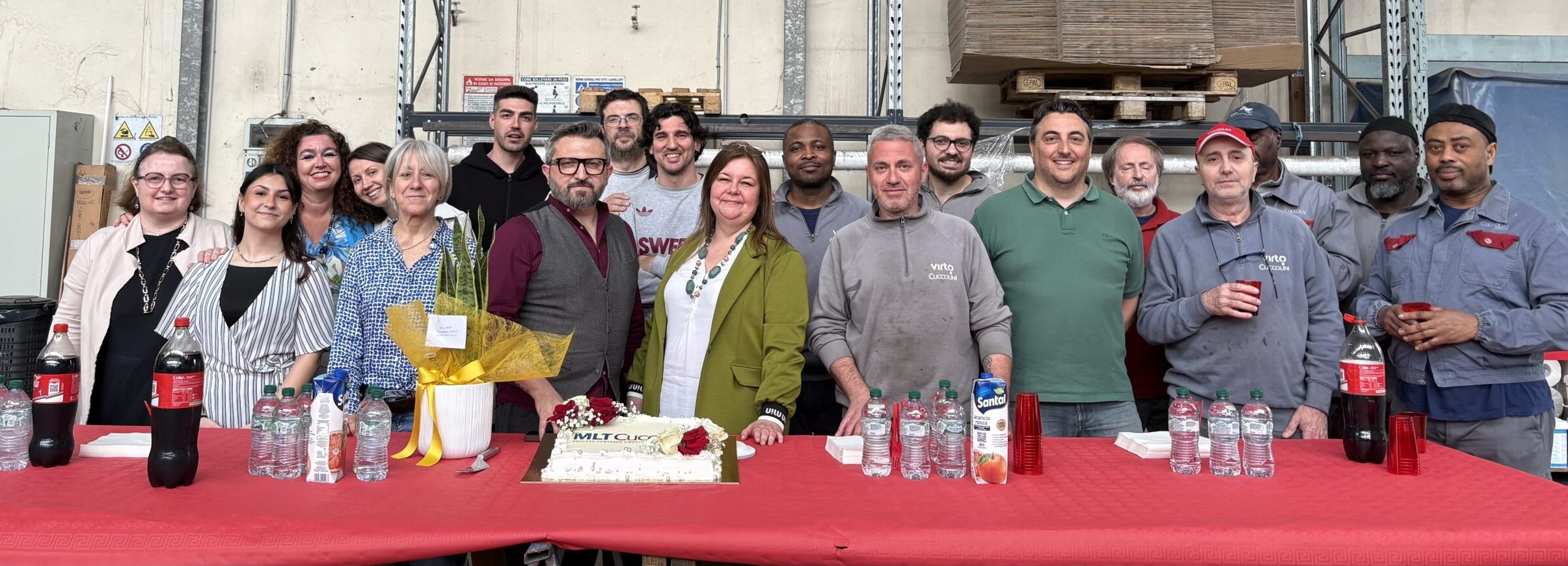 Group photo of about 18 smiling colleagues posing in a warehouse behind a red-covered table with drinks, a decorated cake reading “MLT Cuccolini”. Company celebration scene.